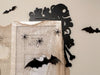 A black door frame topper, designed with the silhouette of scurrying rats, bones, and a skull, is seen on top of a white picture frame that's decorated with fake spiders and spider webs. The beige wall behind has fake bats attached to it.