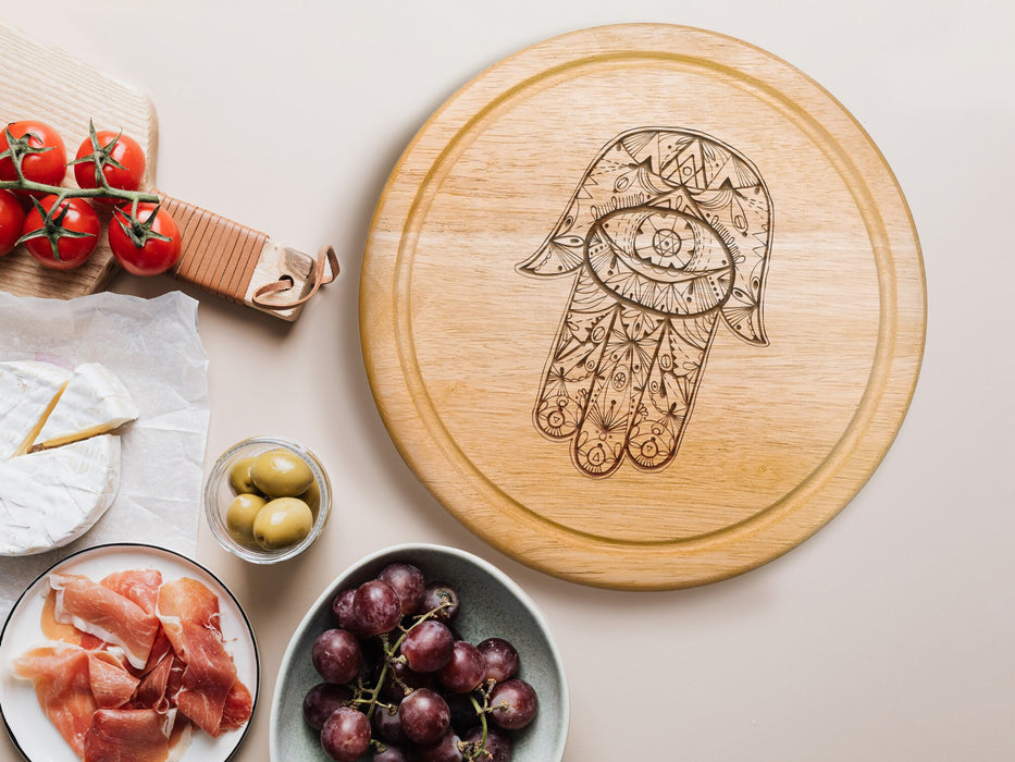 wooden cheeseboard on white table surrounded by charcuterie foods such as Italian meats, olives, grapes, tomatoes and cheese, engraving has a hamsa design