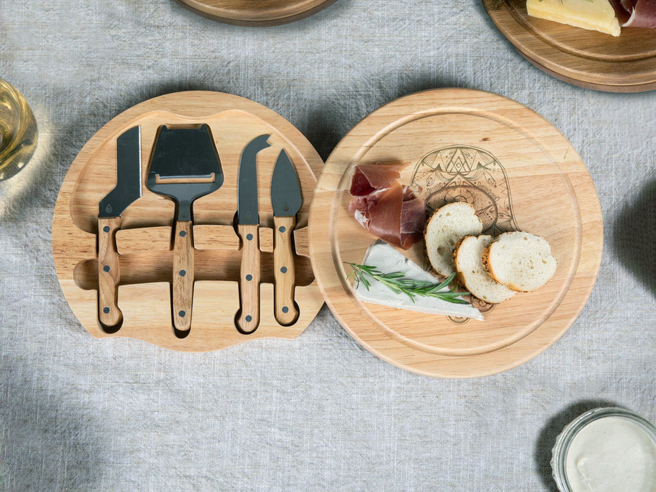 open engraved charcuterie board revealing stainless steel cheese knife set with cheese, meats, and breads ontop surrounded by table cloth, glasses, etc