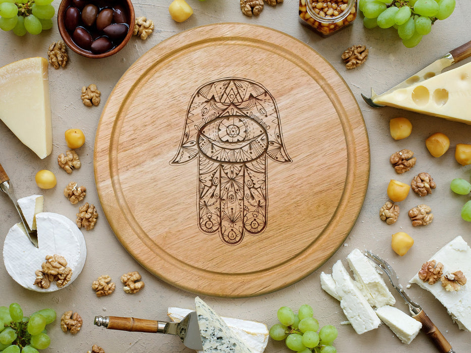 wooden cheese board surrounded by various cheeses, grapes, cheese knife tools, corn nuts, olives and walnuts  engraving is of a hamsa design