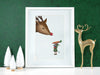 white frame with christmas print of an elf mouse offering a carrot to a reindeer against a green background ontop of a white counter surrounded by holiday decor such as white pine trees and a golden sparkly reindeer
