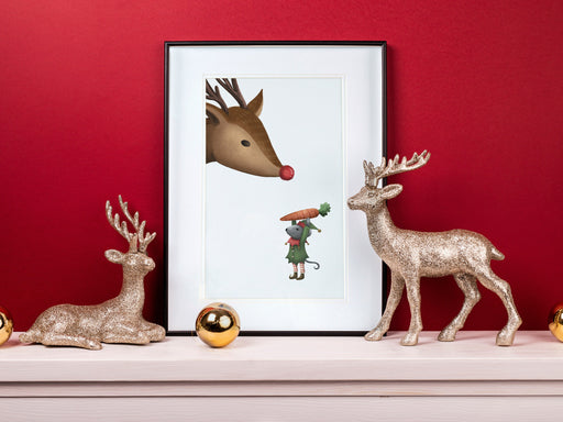 large frame with a christmas print of a mouse elf holding a carrot to a reindeer against a red background ontop of a white wooden countertop surrounded by holiday decor such as golden ornaments and gold sparkly reindeer