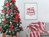 silver frame hanging on white wall with a merry christmas typography print surrounded by holiday decor such as a christmas tree with bows and ornaments, christmas gifts, and a grey couch with a red and white blanket