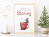 wooden frame hanging over wooden counter with christmas print of a mouse stirring hot chocolate with marshmallows and typography surrounded by holiday decor such as white stars, mini wooden houses, and wrapped gifts