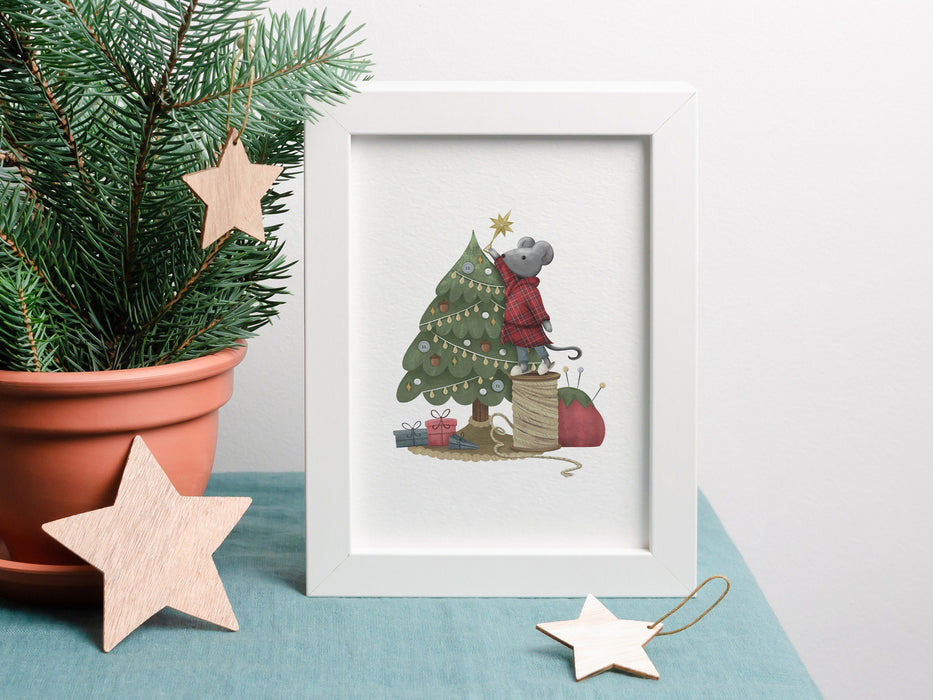 small Christmas print of mouse decorating tree inside a white frame ontop of a blue table cloth next to a mini pine tree surrounded by wooden stars