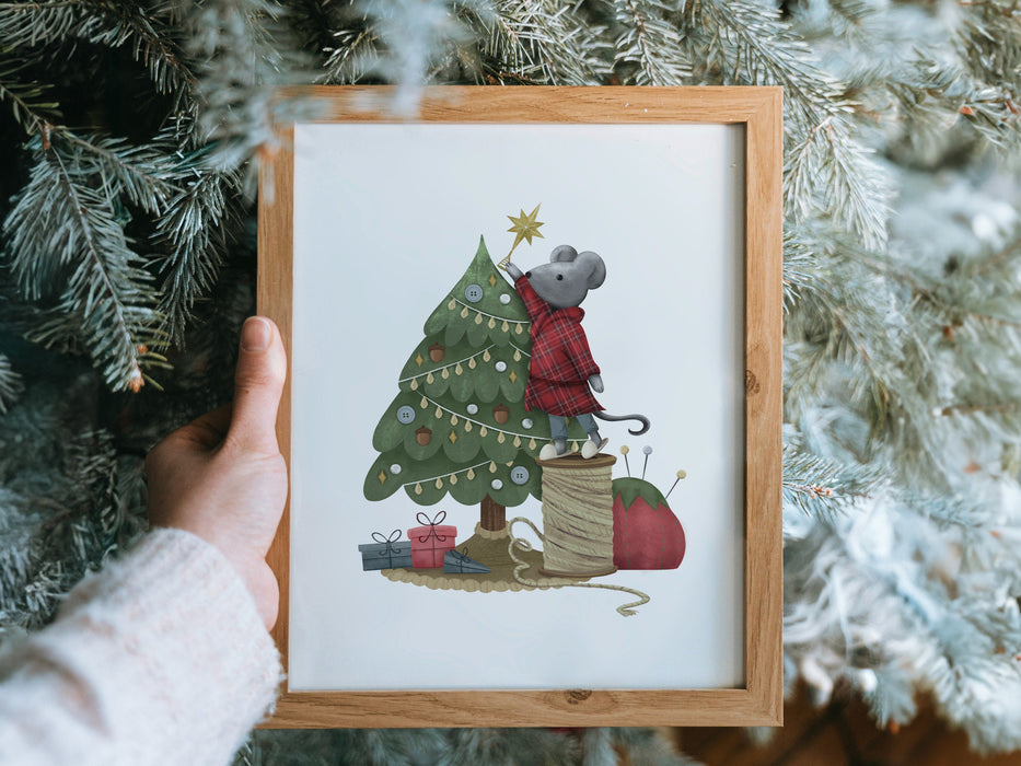 hand holding a wooden frame with a christmas print of a mouse decorating a tree in front of pine trees