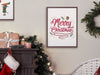 brown frame with merry christmas typography print on white wall surrounded by holiday decor such as pine leaves, christmas lights, stocking, christmas gift, white chair and pillow, next to a brown fireplace