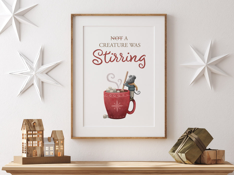 wooden frame hanging over wooden counter with christmas print of a mouse stirring hot chocolate with marshmallows and typography, surrounded by holiday decor such as white stars, mini wooden houses, and wrapped gifts