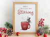 wooden frame with christmas print of mouse stirring hot chocolate with marshmallows and typography ontop of wooden countertop surrounded by winter decor such as pinecones, pine leaves, holly berries, and red ornaments