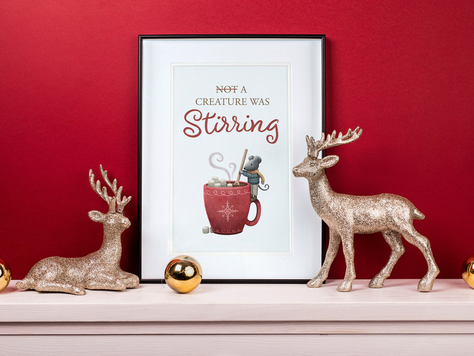 large frame with a christmas print of a mouse stirring hot chocolate with marshmallows and typography against a red background ontop of a white wooden countertop surrounded by holiday decor such as golden ornaments and gold sparkly reindeer