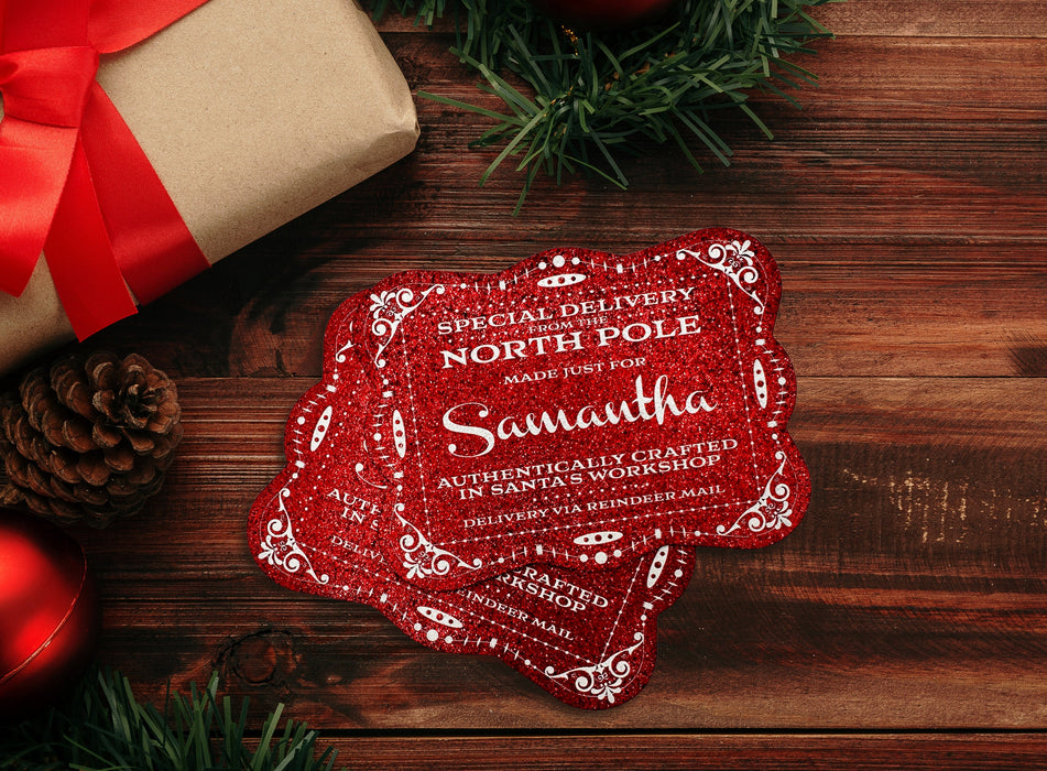 Two red glitter cardstock Santa gift tags are shown on a dark wooden surface. Pine tree branches, a pine cone, a red ornament, and a Christmas present are shown around the tags.