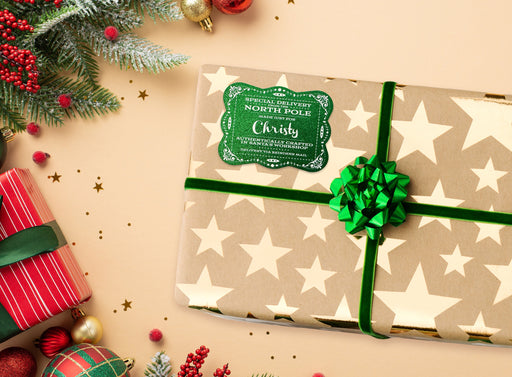 A green glitter cardstock Santa gift tag is shown on a Christmas present. The present paper is made of craft paper and has gold stars on it. The box is seen on a tan surface with other Christmas elements around it.