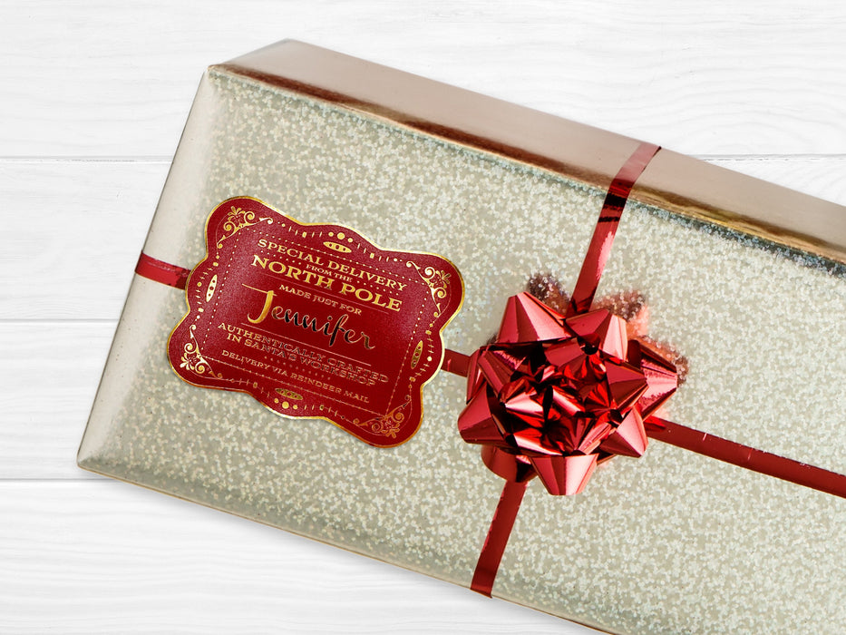 A gold foil cardstock Santa gift tag is shown on a Christmas present. The present is wrapped in gold paper and has a red bow and ribbon around it. The background of the picture is a white wood surface.