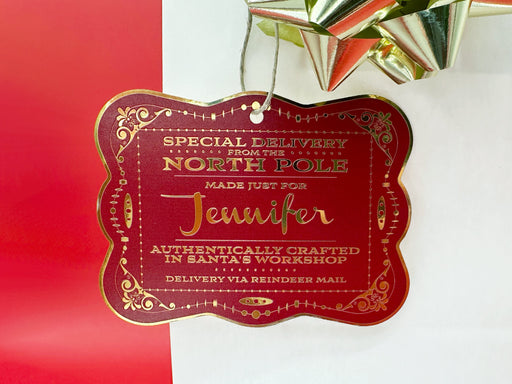 A gold foil cardstock Santa gift tag is shown hanging on a white gift bag. The bag is on a red backdrop and has a gold bow on it.