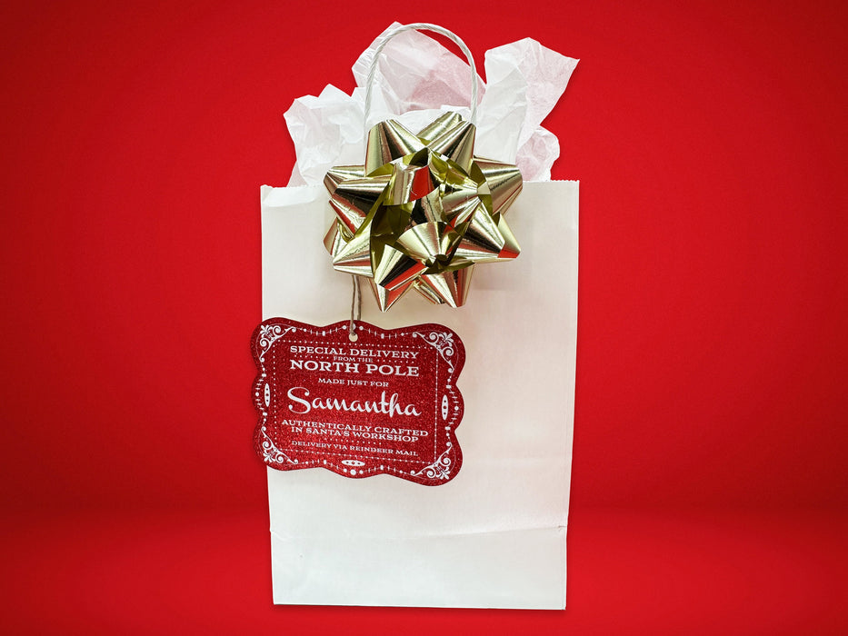A red glitter cardstock Santa gift tag is shown hanging on a white gift bag. The bag is on a red backdrop and has a gold bow on it.