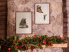 two wooden framed christmas prints hung on stone hanging over christmas decor such as holly berry, candles, and pine leaves