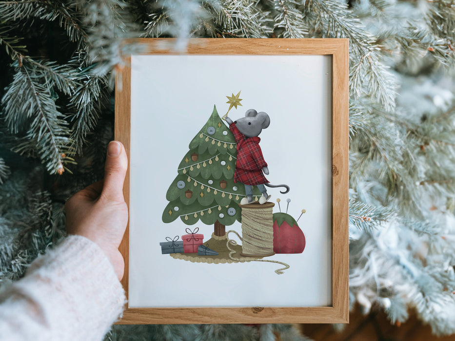 hand holding a wooden frame with christmas print of mouse decorating christmas tree in front of pine trees