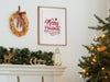 wooden frame hanging on wall over a fireplace with merry christmas typography print surrounded by holiday decor such as pine leaves, christmas tree, christmas lights, wooden cutouts of reindeer and sleigh, rustic decor