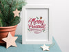 small merry christmas typography print inside a white frame ontop of a blue table cloth next to a mini pine tree surrounded by wooden stars