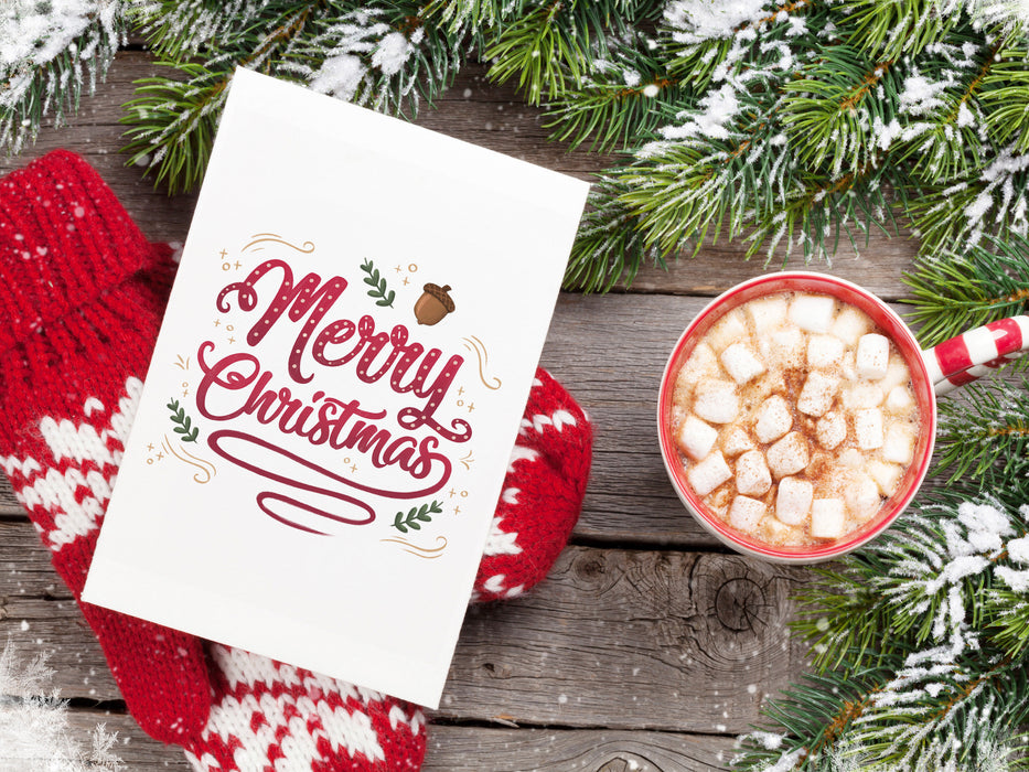 wooden table with merry christmas typography print, red and white gloves, and hot cocoa with marshmallows ontop surrounded by pine leaves covered in snow