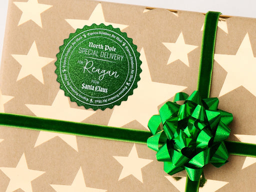 A green glitter cardstock Santa gift tag is shown on a Christmas present. The present paper is made of craft paper and has gold stars on it. The box is seen on a white surface.