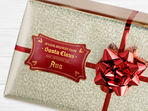 A gold foil cardstock Santa gift tag is shown on a Christmas present. The present is wrapped in gold paper and has a red bow and ribbon around it. The background of the picture is a white wood surface.