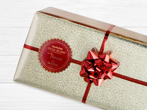 A gold foil cardstock Santa gift tag is shown on a craft paper wrapped box. The box also has a red and white striped string on it. The box is seen on a wooden surface.