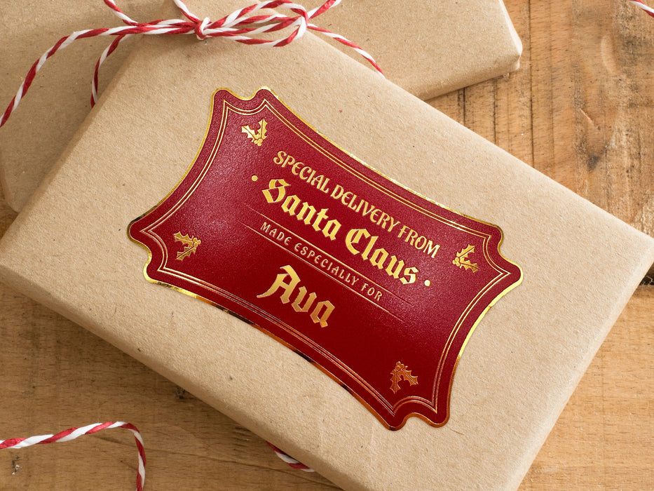 A gold foil cardstock Santa gift tag is shown on a craft paper wrapped box. The box also has a red and white striped string on it. The box is seen on a wooden surface.