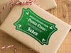 A green glitter cardstock Santa gift tag is shown on a craft paper wrapped box. The box also has a red and white striped string on it. The box is seen on a wooden surface.