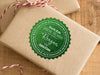 A green glitter cardstock Santa gift tag is shown on a craft paper wrapped box. The box also has a red and white striped string on it. The box is seen on a wooden surface.