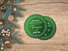 Two green glitter cardstock Santa gift tags are shown on a wooden table. The table has gold Christmas ornaments and pine tree branches on it.