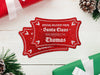 Two red glitter cardstock Santa gift tags are shown on a white wooden surface. Pine tree branches, pine cones, and Christmas presents are shown around the tags.