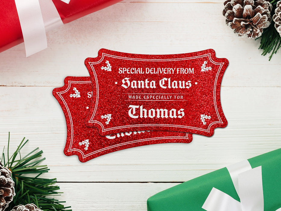 Two red glitter cardstock Santa gift tags are shown on a white wooden surface. Pine tree branches, pine cones, and Christmas presents are shown around the tags.