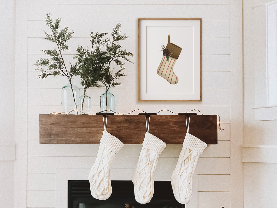 wooden frame with christmas print of a rustic stocking hanging on wall over fireplace with potted house plants and white knitted stockings