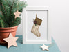 small Christmas print of a rustic stocking inside a white frame ontop of a blue table cloth next to a mini pine tree surrounded by wooden stars