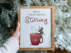hand holding a wooden frame with a christmas print of a mouse stirring hot chocolate with typography in front of pine trees
