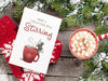 wooden table with christmas print of a mouse stirring hot chocolate with typography, red and white gloves, and hot cocoa with marshmallows ontop surrounded by pine leaves covered in snow