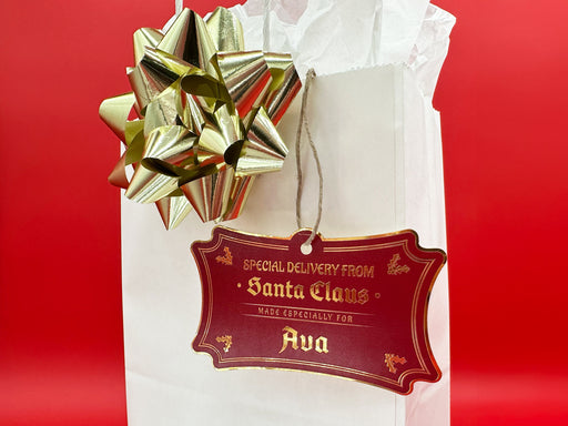 A gold foil cardstock Santa gift tag is shown hanging on a white gift bag. The bag is on a red backdrop and has a gold bow on it.