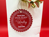 A red glitter cardstock Santa gift tag is shown hanging on a white gift bag. The bag is on a red backdrop and has a gold bow on it.