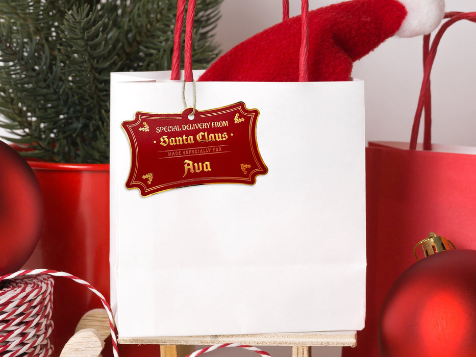 A gold foil cardstock Santa gift tag is shown hanging on a white gift bag. The bag is sitting on a sled and is surrounded by Christmas ornaments, a Christmas tree, and a red gift bag. Everything is set against a white backdrop.