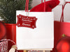 A red glitter cardstock Santa gift tag is shown hanging on a white gift bag. The bag is sitting on a sled and is surrounded by Christmas ornaments, a Christmas tree, and a red gift bag. Everything is set against a white backdrop.