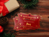 Two gold foil cardstock Santa gift tags are shown on a dark wooden surface. A pine cone, pine branches,  a red Christmas ornament, and a gift box can be seen on the surface by the tags.