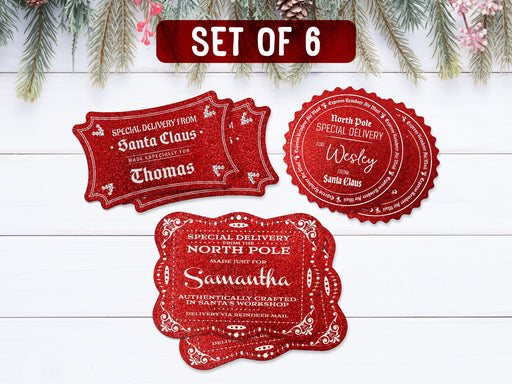 Text reads, Set of 6. Six red glitter cardstock Santa gift tags are shown on a white wooden surface. Pine tree branches, pine cones, and other foliage can be seen on the top of the image.