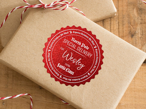 A red glitter cardstock Santa gift tag is shown on a craft paper wrapped box. The box also has a red and white striped string on it. The box is seen on a wooden surface.
