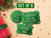Text reads, Set of 6. Three green glitter cardstock Santa gift tags are shown on a tan background. Pine tree branches, holly, Christmas ornaments, gold glitter, and a red and green gift-wrapped box can also be seen.