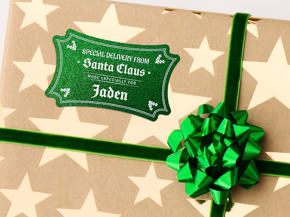 A green glitter cardstock Santa gift tag is shown on a Christmas present. The present paper is made of craft paper and has gold stars on it. The present is shown on a white background.