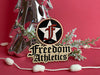 Custom ornament for Freedom Athletics is shown. Ornament is laser cut to custom shape and includes business logo. Ornament is shown against a red background with a silver Christmas tree behind it with pine branches.