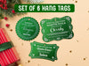 Text reads, Set of 6 Hang Tags. Three green glitter cardstock hanging Santa gift tags are shown on a tan background. Pine tree branches, holly, Christmas ornaments, gold glitter, and a red and green gift-wrapped box can also be seen.