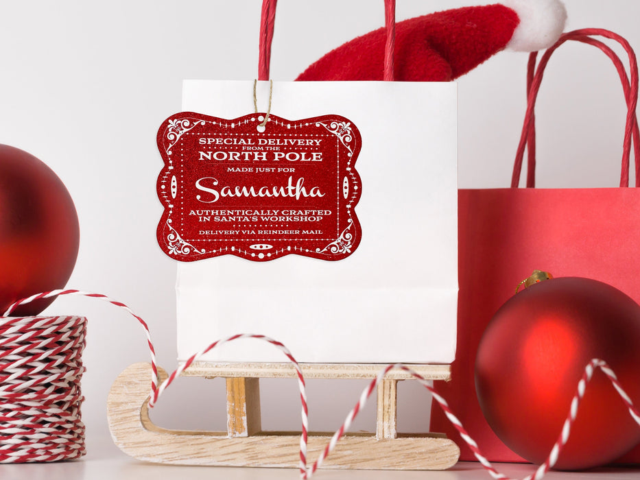 A red glitter cardstock Santa gift tag is shown hanging on a white gift bag. The bag is sitting on a sled and is surrounded by Christmas ornaments, a Santa hat, and a red gift bag. Everything is set against a white backdrop.