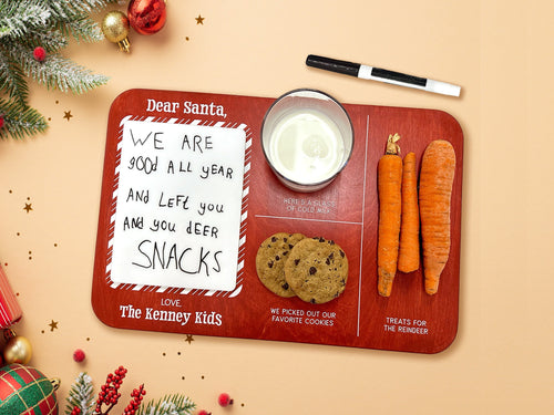 red Santa cookies and milk tray with a glass of milk, cookies, and carrots ontop of a beige background next to a dry erase marker surrounded by holiday decor such as pine leaves, holly berries, and christmas ornaments